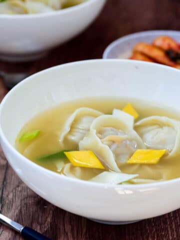 Korean dumpling soup in a white bowl garnished with small diamond shape egg crepe served with a side of kimchi