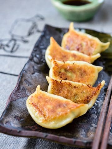 5 pan-fried shrimp dumplings on a dark stone plate with chopsticks on and a dipping sauce at the top